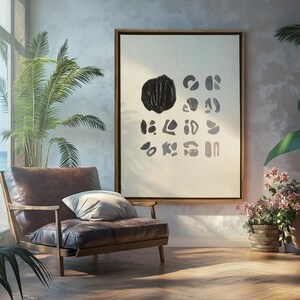 May include: A framed abstract art print with black and white shapes on a white background. The print is hanging on a light gray wall in a room with a brown leather armchair and plants.