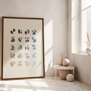 May include: A framed abstract art print featuring a series of blue and gray organic shapes on a cream background. The artwork is displayed in a light-colored room with a small side table and a window.