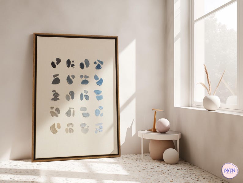 May include: A framed abstract art print featuring a series of blue and gray organic shapes on a cream background. The artwork is displayed in a light-colored room with a small side table and a window.