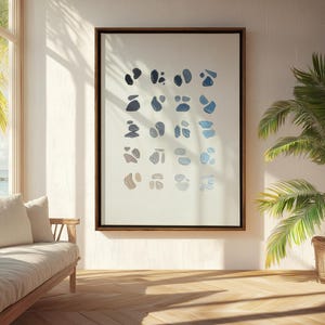 May include: A framed abstract art print featuring a series of blue and gray organic shapes on a white background. The artwork is displayed in a room with a wooden floor, a sofa, and a palm tree in a woven basket.