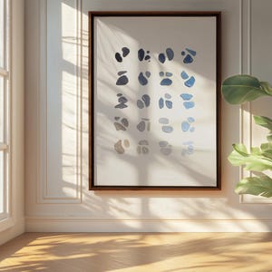 May include: A framed abstract art print featuring a series of blue and gray organic shapes on a white background. The artwork is displayed in a room with natural light, a large window, and a potted plant.