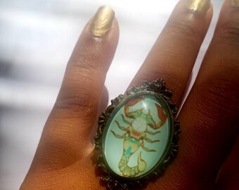 May include: A ring with a bronze-colored setting and a glass cameo featuring a detailed illustration of a scorpion in shades of green, yellow, and orange.