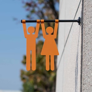 Op de afbeelding: Oranje silhouetten van twee personen, een met een rok, hangend aan een zwarte metalen balk. De figuren zijn met metalen haken aan de balk bevestigd.