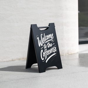 May include: A black A-frame sign with white lettering reading "Welcome to the Coffeeverse". The sign, with a handle, is positioned on a grey surface. It's a promotional sign, likely for a coffee shop or cafe, designed to attract customers.