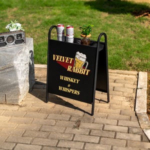 May include: Black metal A-frame sign with gold lettering reading "Velvet Rabbit Whiskey & Whispers". Two Budweiser cans and a small plant are on the top shelf. The sign is on a brick patio outdoors.