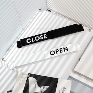 May include: A black sign with the word "CLOSE" in white, and a white sign with "OPEN" in black. The signs are on a white surface, with a magazine and a book visible in the background. The signs are likely for a shop.