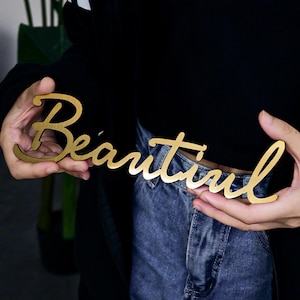 Puede incluir: Un letrero de madera de color dorado con la palabra "Beautiful" escrita en letra cursiva.