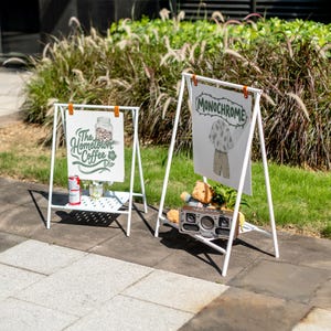 May include: Two white A-frame signs with printed graphics. One sign reads "The Hometown Coffee" with a coffee bean jar illustration. The other sign says "MONOCHROME" with a clothing illustration. A small shelf holds items.