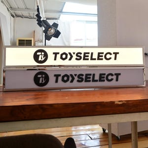 May include: Two white rectangular signs with black lettering. The top sign is lit up and the bottom sign is not. Both signs say "Ti TOYSELECT".