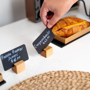 Op de afbeelding: Een hand plaatst een klein zwart schoolbordje met de tekst "Croquembouche €23,00" in een houten houder. Een croissant sandwich in een kartonnen bakje staat op de achtergrond. Een ander bordje luidt "Raisin Butter Scones €16,00".