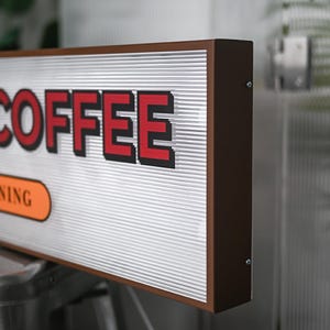 Könnte beinhalten: Ein braunes rechteckiges Schild mit dem Wort "COFFEE" in roten Buchstaben mit schwarzem Umriss. Das Schild hat eine weiße, wellenförmige Kunststoffoberfläche und einen braunen Rahmen.