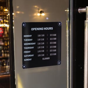 以下が含まれることがあります： ガラスドアに取り付けられた、営業時間を表示する黒い看板。白い文字で「OPENING HOURS」と書かれており、曜日と対応する時間が表示されています。看板は4つの銀色の留め具で固定されています。