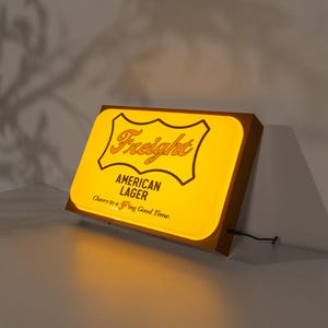 以下が含まれることがあります： 「Freight American Lager Cheers to a  'Fring Good Time.」と書かれた黄色と茶色の照明付き看板