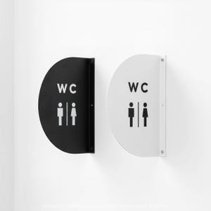May include: Two semi-circular wall-mounted signs, one black and one white, each featuring the text WC and a graphic icon of a person in a dress and a person in trousers separated by a vertical line. The signs have a flat mounting bracket.