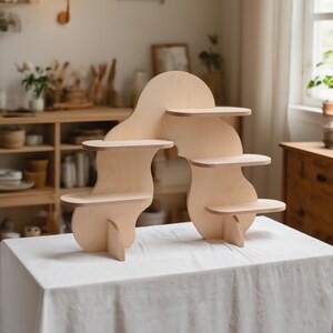 May include: A light-colored wooden display shelf with a unique, curved design. It features multiple small, round shelves of varying heights, supported by a flowing, organic frame. The shelf is placed on a white tablecloth.