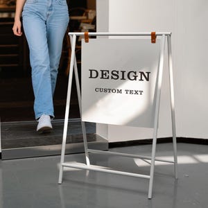 Pode incluir: Um sinal branco em forma de A com o texto "DESIGN CUSTOM TEXT" em um fundo branco. O sinal é feito de metal e tem tiras de couro marrom.