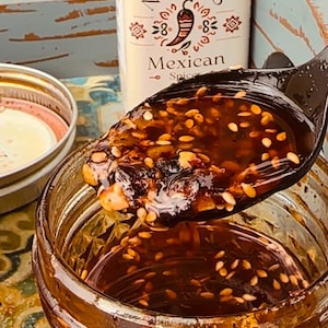 May include: A close-up of a jar filled with a dark, glistening sauce, with a spoonful lifted above it. The sauce is speckled with sesame seeds and other visible spices. A bottle labeled "Nena's Mexican Spice" is in the background.