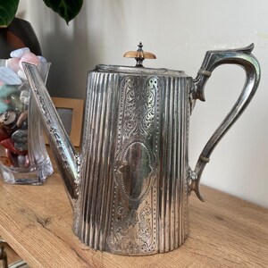 May include: A silver teapot with an ornate design and a wooden knob on the lid. The teapot has a tall, cylindrical body and a curved spout.