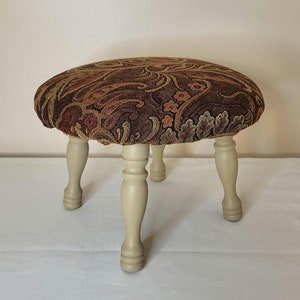 Puede incluir: Un pequeño taburete redondo con estampado floral y cuatro patas de madera torneadas. El taburete está pintado de blanco y tiene un estampado floral marrón, naranja y dorado.
