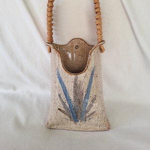 May include: A ceramic wall vase with a woven wicker handle. The vase is decorated with a blue and brown floral design on a speckled beige background.