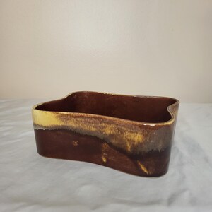 May include: A brown ceramic planter with a unique wavy design. The planter has a dark brown base that fades to a lighter brown and yellow rim. The open top is rectangular.