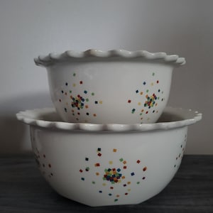 May include: Two white ceramic bowls stacked, each with a scalloped rim. The bowls are decorated with scattered, colorful square confetti. The bowls are on a dark gray surface.