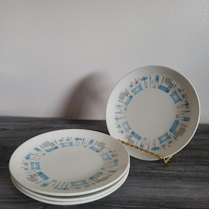 Op de afbeelding: Een set van vier witte keramische borden met een modern design uit het midden van de eeuw. Elk bord heeft een herhalend patroon van blauwe en grijze geometrische vormen rond de rand. Eén bord wordt op een goudkleurige standaard gepresenteerd.