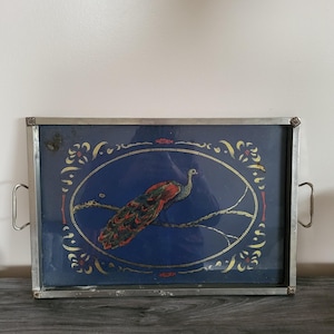 May include: A rectangular serving tray with a metallic silver frame and handles. The center features a deep blue background with a central oval border containing a colorful peacock perched on a branch, accented by decorative gold floral motifs.