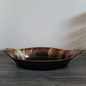 Peut inclure: Un plat de service en céramique noire avec un bord marron et beige. Le plat a un bord ondulé avec des poignées de chaque côté. L'intérieur du plat est noir et l'extérieur est brillant.