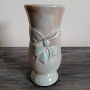 May include: A ceramic vase with a bow design. The vase has a light blue and peach color scheme. The bow is a lighter shade of blue. The vase has a pedestal base and a flared top.