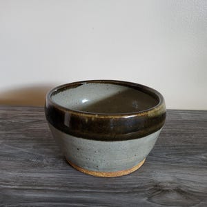 May include: A handcrafted ceramic bowl with a grey and brown glaze. The bowl has a rounded form with a dark brown band around the middle and a light grey interior. The base is a natural, unglazed colour.