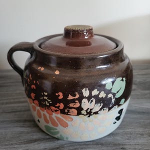 Puede incluir: Un tarro de cerámica marrón con tapa y asa. El tarro está decorado con un estampado floral en tonos naranja, rosa y verde sobre una base blanca. La tapa es de color marrón oscuro. Capacidad aproximada de 470 ml.