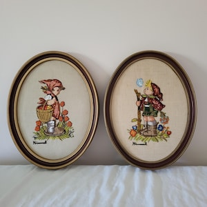 May include: Two framed oval needlepoint artworks. One depicts a girl with a basket of fruit, the other a boy with a stick and a bubble. Both are set against a beige background, framed in brown and gold, with the word "Hummel" at the bottom.