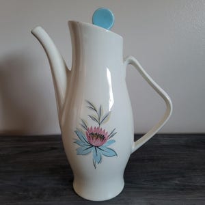 Puede incluir: Una cafetera de cerámica blanca con un pico largo y un asa. La cafetera presenta un diseño floral en rosa, azul y verde. Una tapa azul claro se encuentra en la parte superior. La cafetera está sobre una superficie de madera oscura.