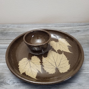 May include: A brown ceramic serving dish with a small bowl in the center. The dish features three beige leaf designs. The dish is designed for serving snacks or appetizers.