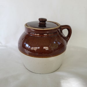 Puede incluir: Un tarro de cerámica esmaltado en marrón y blanco con tapa y asa. El tarro es de estilo tradicional con cuerpo redondeado y boca ancha.