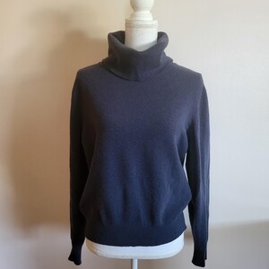 Puede incluir: Un jersey de cuello alto azul marino exhibido en un maniquí blanco. El jersey tiene mangas largas y un cuello alto y doblado. La tela parece suave y ligeramente texturizada, con un brillo sutil. El fondo es una pared lisa de color claro.