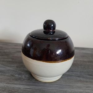 Puede incluir: Un azucarero redondo de cerámica con tapa. La mitad superior del cuenco y la tapa son de color marrón oscuro, mientras que la mitad inferior es de color crema. Una fina banda dorada separa los dos colores. La tapa tiene un pequeño pomo redondo.