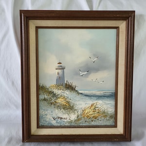 Puede incluir: Una pintura enmarcada de un faro blanco en una playa de arena con gaviotas volando por encima. La pintura está realizada en un estilo realista con colores apagados.