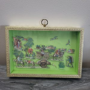 Pode incluir: Uma caixa de sombra emoldurada com uma moldura dourada ornamentada. O interior apresenta um fundo verde com um diorama detalhado retratando figuras, cavalos e uma carruagem em uma cena pastoral. Um pequeno anel de latão está no topo.