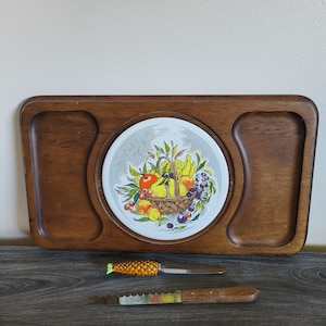 May include: A vintage wooden serving tray with a round ceramic insert featuring a fruit basket design. Includes two knives, one with a pineapple-shaped handle. The tray has two recessed areas and a dark wood finish.