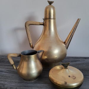 May include: A vintage brass tea set comprising a tall teapot with a wooden handle, a small creamer, and a lidded sugar bowl. The set has a warm, golden colour and a classic design, ideal for serving tea or coffee.