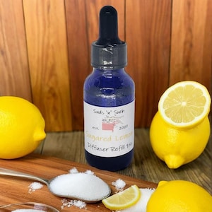 May include: A blue glass bottle with a dropper top, labeled "Suds 'n Such est. 2007 Sugared Lemon Diffuser Refill 1oz". The bottle is surrounded by lemons and a spoon with white sugar crystals.