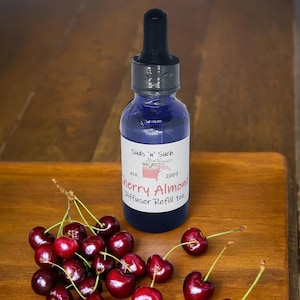 May include: A blue glass bottle with a dropper top, labeled "Suds 'n' Such est. 2007 Cherry Almond Diffuser Refill 1oz". The bottle is sitting on a wooden surface with a group of red cherries.