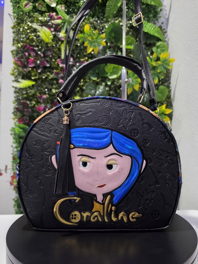 May include: A black dome-shaped handbag featuring an embossed pattern of keys and buttons. The front displays a raised, colorful character graphic of Coraline with blue hair and the name Coraline in gold lettering. It includes a black tassel charm.