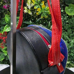 May include: A round crossbody bag featuring a black textured exterior with a red zipper closure and matching red straps. The side panel displays a vibrant blue border with a subtle iridescent finish, and the bag is suspended from a metal display stand.