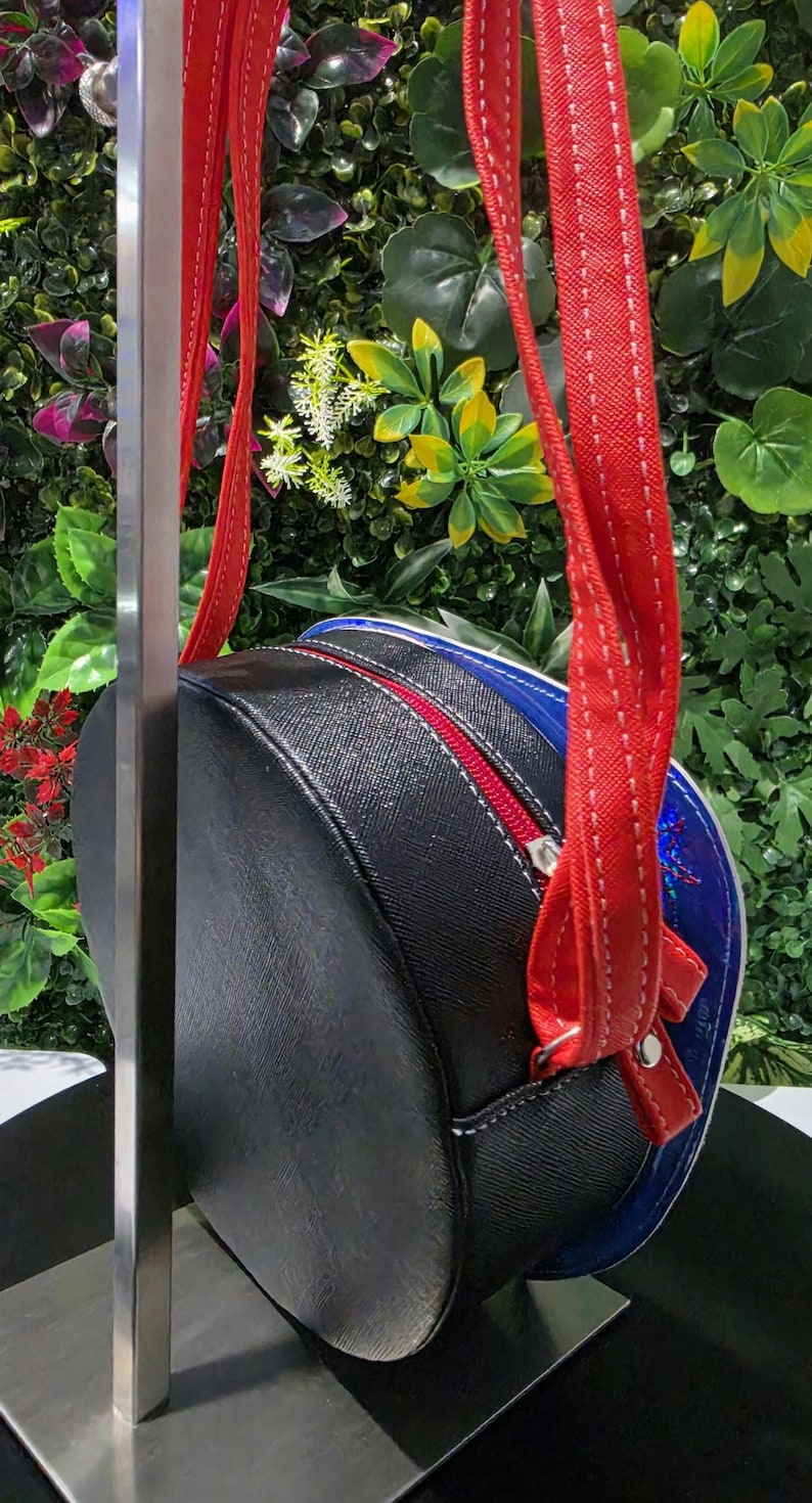May include: A round crossbody bag featuring a black textured exterior with a red zipper closure and matching red straps. The side panel displays a vibrant blue border with a subtle iridescent finish, and the bag is suspended from a metal display stand.