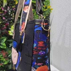 May include: A black tote bag featuring a colorful side panel with a graphic print of a black cat, a key, and a pumpkin with the text Welcome home. The bag has a patterned shoulder strap, a black tassel, and a cartoon character on the front panel.