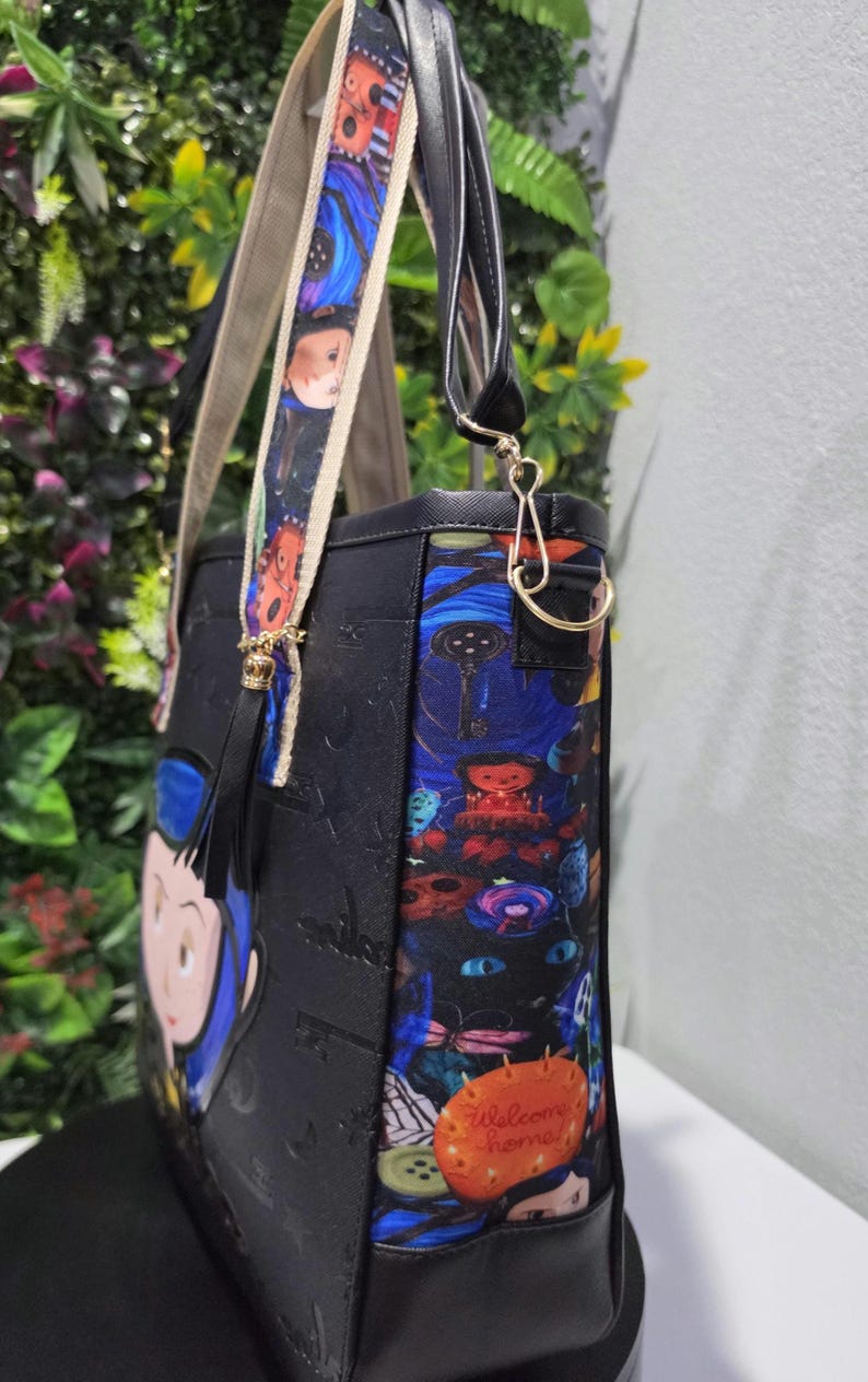 May include: A black tote bag featuring a colorful side panel with a graphic print of a black cat, a key, and a pumpkin with the text Welcome home. The bag has a patterned shoulder strap, a black tassel, and a cartoon character on the front panel.