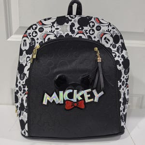 May include: A black and white backpack featuring a repeating Mickey Mouse pattern on the sides and top. The front pocket is solid black with a debossed Mickey icon pattern, a holographic MICKEY text logo, and a red bow tie accent.